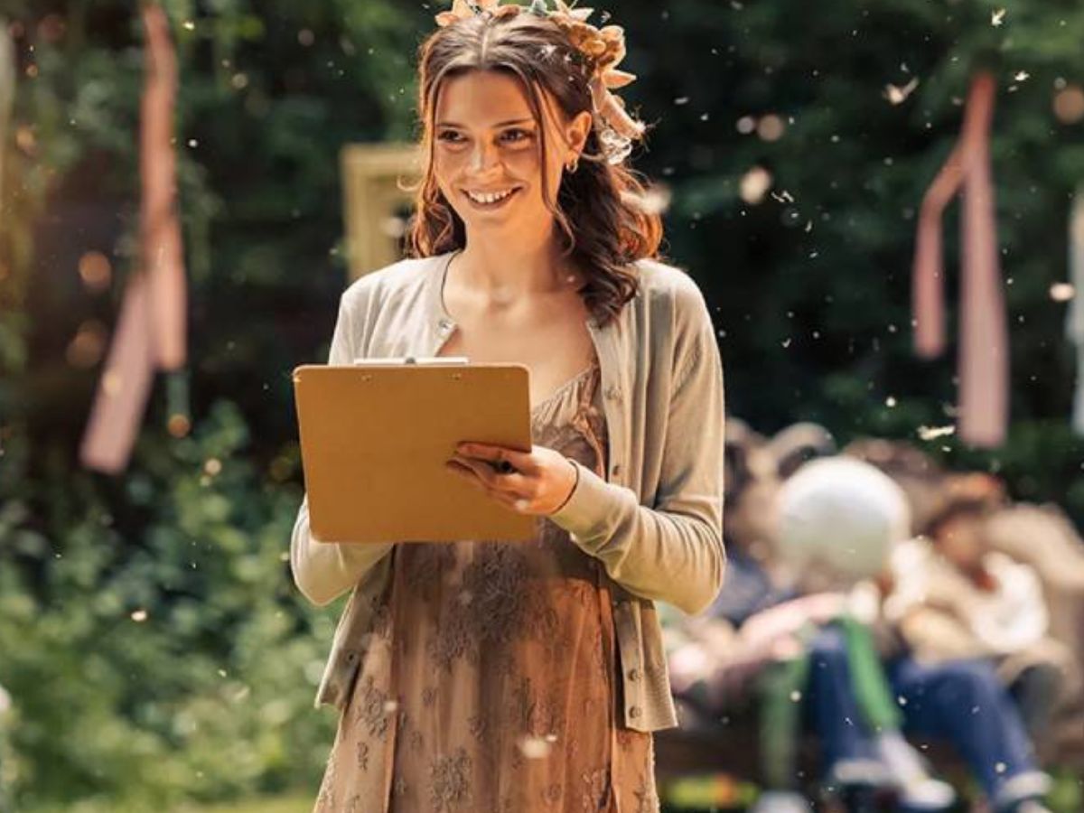 Na foto há uma mulher usando uma coroa de flores e segurando uma prancheta. Ela está sorrindo.