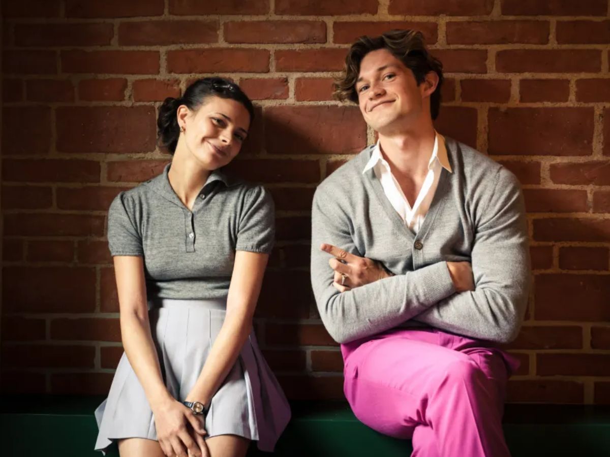 Na foto há uma mulher usando uma blusa polo cinza e uma saia cinza. Ela está sentada encostada em uma parede de tijolos. Há também um homem sentado do mesmo jeito e usando um casaco cinza e uma calça rosa.