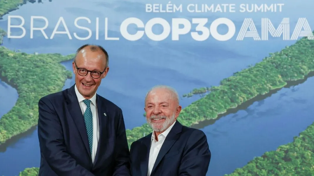 A imagem registra um encontro entre o presidente do Brasil e o líder alemão. O chanceler alemão, Friedrich Merz, está de pé, voltado para a frente. Ele usa óculos e está vestido com um terno azul-marinho escuro e uma gravata azul-clara estampada. Ele sorri de forma aberta, virando levemente a cabeça em direção ao seu colega brasileiro. O presidente brasileiro, Luiz Inácio Lula da Silva, está de pé ao lado dele, também sorrindo amplamente e olhando para a frente. Ele veste um terno escuro sobre uma camisa social branca de colarinho aberto (sem gravata) e tem barba e cabelo grisalhos.