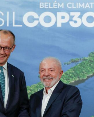 A imagem registra um encontro entre o presidente do Brasil e o líder alemão. O chanceler alemão, Friedrich Merz, está de pé, voltado para a frente. Ele usa óculos e está vestido com um terno azul-marinho escuro e uma gravata azul-clara estampada. Ele sorri de forma aberta, virando levemente a cabeça em direção ao seu colega brasileiro. O presidente brasileiro, Luiz Inácio Lula da Silva, está de pé ao lado dele, também sorrindo amplamente e olhando para a frente. Ele veste um terno escuro sobre uma camisa social branca de colarinho aberto (sem gravata) e tem barba e cabelo grisalhos.