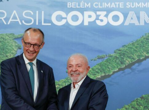 A imagem registra um encontro entre o presidente do Brasil e o líder alemão. O chanceler alemão, Friedrich Merz, está de pé, voltado para a frente. Ele usa óculos e está vestido com um terno azul-marinho escuro e uma gravata azul-clara estampada. Ele sorri de forma aberta, virando levemente a cabeça em direção ao seu colega brasileiro. O presidente brasileiro, Luiz Inácio Lula da Silva, está de pé ao lado dele, também sorrindo amplamente e olhando para a frente. Ele veste um terno escuro sobre uma camisa social branca de colarinho aberto (sem gravata) e tem barba e cabelo grisalhos.