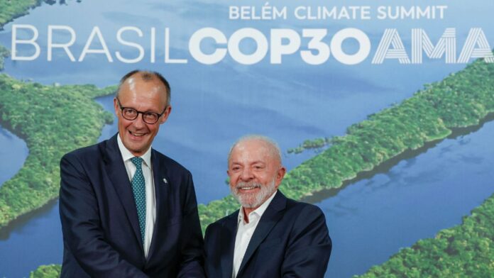 A imagem registra um encontro entre o presidente do Brasil e o líder alemão. O chanceler alemão, Friedrich Merz, está de pé, voltado para a frente. Ele usa óculos e está vestido com um terno azul-marinho escuro e uma gravata azul-clara estampada. Ele sorri de forma aberta, virando levemente a cabeça em direção ao seu colega brasileiro. O presidente brasileiro, Luiz Inácio Lula da Silva, está de pé ao lado dele, também sorrindo amplamente e olhando para a frente. Ele veste um terno escuro sobre uma camisa social branca de colarinho aberto (sem gravata) e tem barba e cabelo grisalhos.