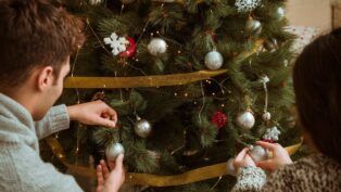 Casal colocando enfeites cinzas em uma grande árvore de Natal verde