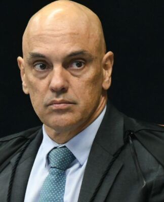 A foto é um retrato em plano médio-fechado, focado no Ministro do Supremo Tribunal Federal (STF), Alexandre de Moraes. O ministro aparece em uma pose formal, olhando diretamente para a frente ou ligeiramente para o lado direito do observador. Ele tem a cabeça raspada/calva e as sobrancelhas arqueadas, com uma expressão séria, tensa e de intensa concentração, que pode ser interpretada como desconfiança ou atenção plena. Seus olhos estão arregalados e fixos.