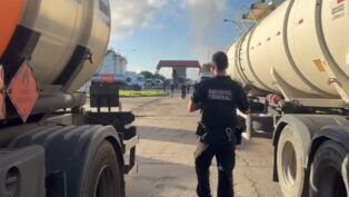 Esta é uma fotografia em plano médio, tirada ao nível do chão, em um ambiente industrial ou de pátio de caminhões sob luz natural intensa. O foco está em um agente de segurança em meio a dois caminhões-tanque.