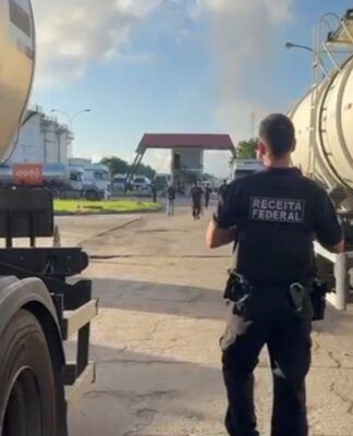 Esta é uma fotografia em plano médio, tirada ao nível do chão, em um ambiente industrial ou de pátio de caminhões sob luz natural intensa. O foco está em um agente de segurança em meio a dois caminhões-tanque.