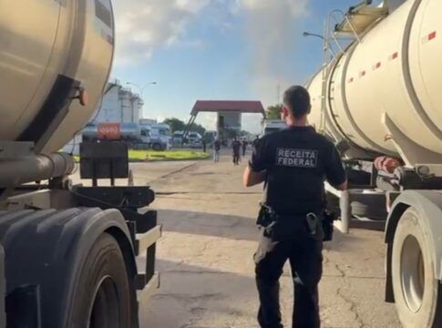 Esta é uma fotografia em plano médio, tirada ao nível do chão, em um ambiente industrial ou de pátio de caminhões sob luz natural intensa. O foco está em um agente de segurança em meio a dois caminhões-tanque.