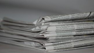 A imagem mostra uma pilha de jornais dobrados, com o papel ligeiramente amassado e textos impressos visíveis nas laterais. Tudo está em tons de cinza, com fundo neutro e desfocado.