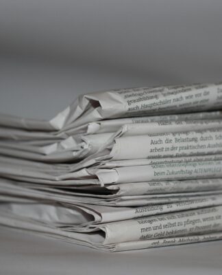 A imagem mostra uma pilha de jornais dobrados, com o papel ligeiramente amassado e textos impressos visíveis nas laterais. Tudo está em tons de cinza, com fundo neutro e desfocado.