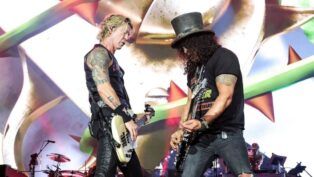 Esta é uma fotografia vibrante tirada no palco durante um show da banda de rock Guns N' Roses. O foco está em dois membros da banda em um momento de performance intensa.