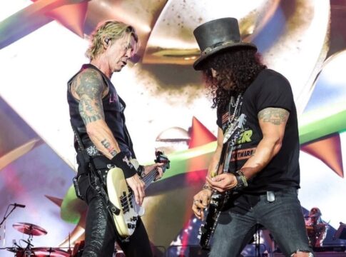Esta é uma fotografia vibrante tirada no palco durante um show da banda de rock Guns N' Roses. O foco está em dois membros da banda em um momento de performance intensa.