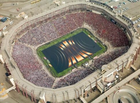 Imagem aérea do Estádio Monumental de Lima completamente lotado durante uma final da Copa Libertadores, com arquibancadas divididas entre torcedores vestidos de vermelho e branco e o gramado coberto por um painel cenográfico com linhas douradas.