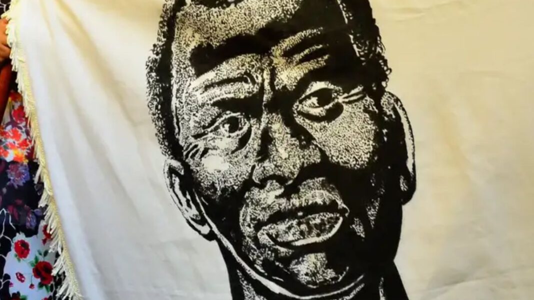 A imagem é um retrato artístico em preto e branco de Zumbi dos Palmares, estampado em um grande tecido ou bandeira de cor creme/branca. O rosto de Zumbi ocupa o centro da imagem. O estilo do desenho é de alto contraste (parecido com xilogravura ou serigrafia), usando densas áreas de preto e branco para criar textura e sombreamento.