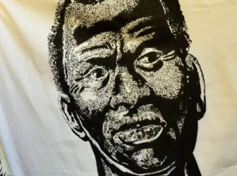 A imagem é um retrato artístico em preto e branco de Zumbi dos Palmares, estampado em um grande tecido ou bandeira de cor creme/branca. O rosto de Zumbi ocupa o centro da imagem. O estilo do desenho é de alto contraste (parecido com xilogravura ou serigrafia), usando densas áreas de preto e branco para criar textura e sombreamento.