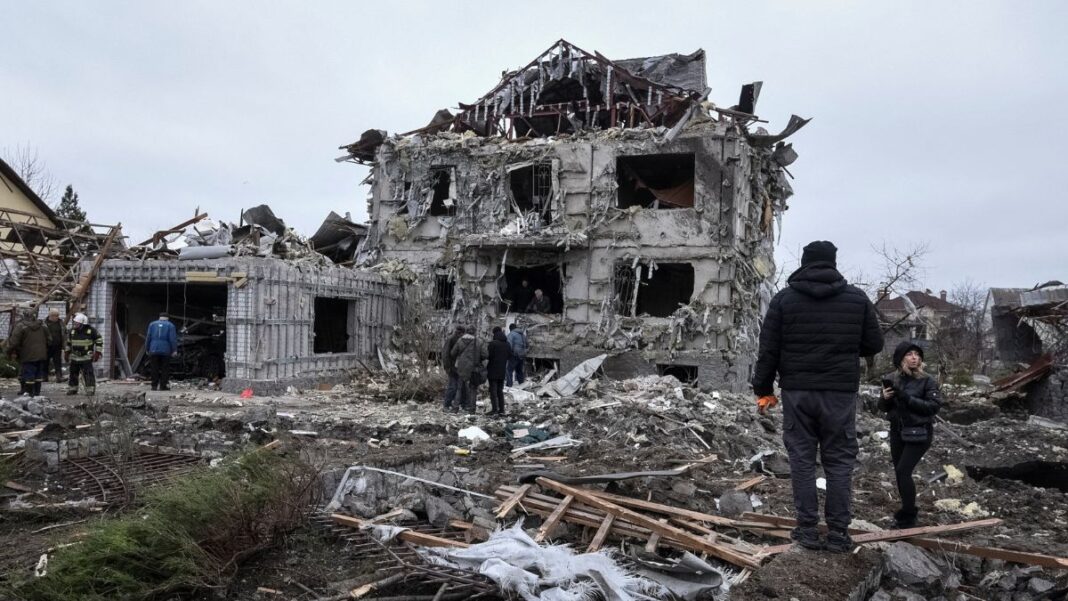 Prédio residencial totalmente destruído, com paredes colapsadas e escombros espalhados por toda a área; várias pessoas caminham e observam os destroços em meio às ruínas, em um cenário cinzento e devastado.