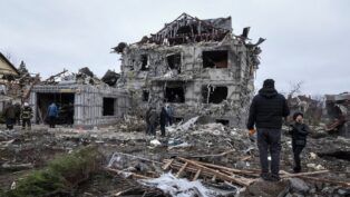 Prédio residencial totalmente destruído, com paredes colapsadas e escombros espalhados por toda a área; várias pessoas caminham e observam os destroços em meio às ruínas, em um cenário cinzento e devastado.