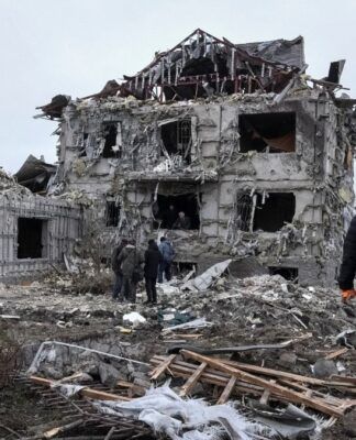 Prédio residencial totalmente destruído, com paredes colapsadas e escombros espalhados por toda a área; várias pessoas caminham e observam os destroços em meio às ruínas, em um cenário cinzento e devastado.