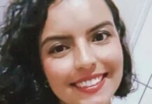 A imagem é um close-up frontal de uma mulher jovem, com expressão sorridente e alegre, tirada em um ambiente interno simples. A mulher está em primeiro plano, ocupando a maior parte da imagem. Ela tem cabelo escuro (castanho ou preto) na altura dos ombros, com cachos volumosos e bem definidos.