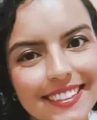 A imagem é um close-up frontal de uma mulher jovem, com expressão sorridente e alegre, tirada em um ambiente interno simples. A mulher está em primeiro plano, ocupando a maior parte da imagem. Ela tem cabelo escuro (castanho ou preto) na altura dos ombros, com cachos volumosos e bem definidos.