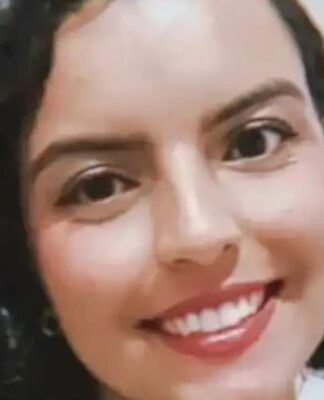 A imagem é um close-up frontal de uma mulher jovem, com expressão sorridente e alegre, tirada em um ambiente interno simples. A mulher está em primeiro plano, ocupando a maior parte da imagem. Ela tem cabelo escuro (castanho ou preto) na altura dos ombros, com cachos volumosos e bem definidos.