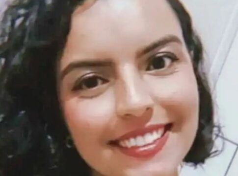 A imagem é um close-up frontal de uma mulher jovem, com expressão sorridente e alegre, tirada em um ambiente interno simples. A mulher está em primeiro plano, ocupando a maior parte da imagem. Ela tem cabelo escuro (castanho ou preto) na altura dos ombros, com cachos volumosos e bem definidos.