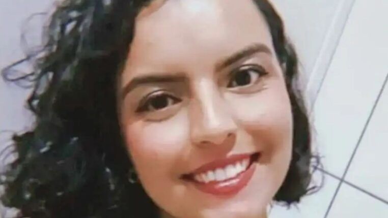 A imagem é um close-up frontal de uma mulher jovem, com expressão sorridente e alegre, tirada em um ambiente interno simples. A mulher está em primeiro plano, ocupando a maior parte da imagem. Ela tem cabelo escuro (castanho ou preto) na altura dos ombros, com cachos volumosos e bem definidos.