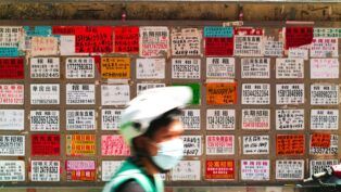 Parede coberta por dezenas de anúncios de aluguel escritos em caracteres chineses, em papéis coloridos e de tamanhos variados; na frente, desfocado, passa um ciclista usando capacete branco, máscara e colete verde.