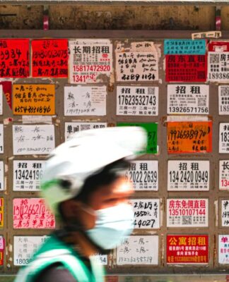 Parede coberta por dezenas de anúncios de aluguel escritos em caracteres chineses, em papéis coloridos e de tamanhos variados; na frente, desfocado, passa um ciclista usando capacete branco, máscara e colete verde.