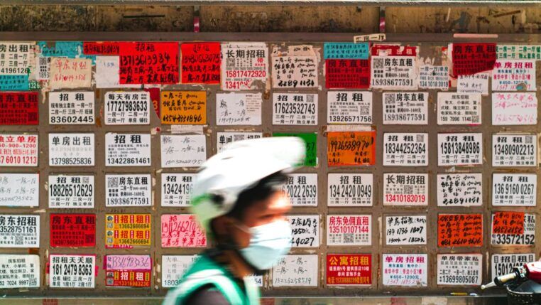 Parede coberta por dezenas de anúncios de aluguel escritos em caracteres chineses, em papéis coloridos e de tamanhos variados; na frente, desfocado, passa um ciclista usando capacete branco, máscara e colete verde.