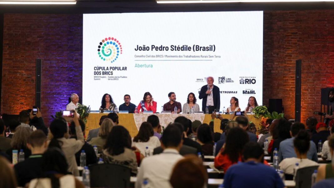 Evento em auditório lotado, com uma mesa de debate composta por vários participantes sentados diante de um grande telão branco. O telão exibe o nome de um palestrante, o logotipo colorido da Cúpula Popular dos BRICS e marcas de patrocinadores. Um dos participantes está de pé falando ao microfone, enquanto o público observa.