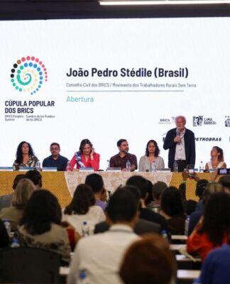 Evento em auditório lotado, com uma mesa de debate composta por vários participantes sentados diante de um grande telão branco. O telão exibe o nome de um palestrante, o logotipo colorido da Cúpula Popular dos BRICS e marcas de patrocinadores. Um dos participantes está de pé falando ao microfone, enquanto o público observa.