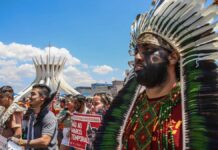 Indígenas marcham em protesto diante da Catedral de Brasília, usando cocares, pinturas corporais e segurando cartazes contra o marco temporal. A cena mostra união, resistência e defesa dos direitos dos povos originários sob um céu ensolarado.