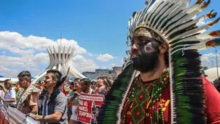 Indígenas marcham em protesto diante da Catedral de Brasília, usando cocares, pinturas corporais e segurando cartazes contra o marco temporal. A cena mostra união, resistência e defesa dos direitos dos povos originários sob um céu ensolarado.