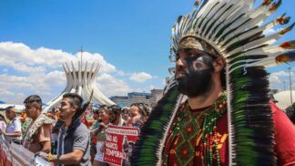 Indígenas marcham em protesto diante da Catedral de Brasília, usando cocares, pinturas corporais e segurando cartazes contra o marco temporal. A cena mostra união, resistência e defesa dos direitos dos povos originários sob um céu ensolarado.