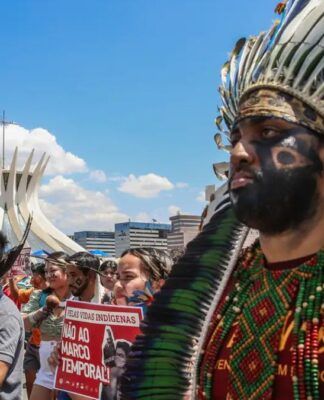 Indígenas marcham em protesto diante da Catedral de Brasília, usando cocares, pinturas corporais e segurando cartazes contra o marco temporal. A cena mostra união, resistência e defesa dos direitos dos povos originários sob um céu ensolarado.