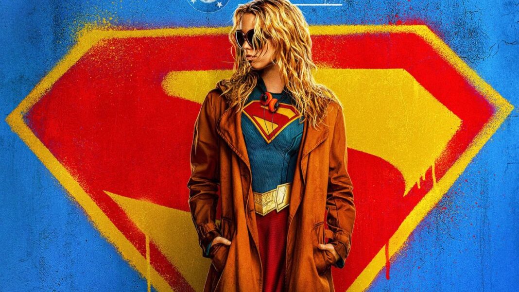 Foto promocional do filme Supergirl onde a atriz Milly Alcock (mulher loira, de óculos escuros e casaco preto por cima do uniforme de super-heroi) aparece olhando para o lado