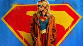 Foto promocional do filme Supergirl onde a atriz Milly Alcock (mulher loira, de óculos escuros e casaco preto por cima do uniforme de super-heroi) aparece olhando para o lado