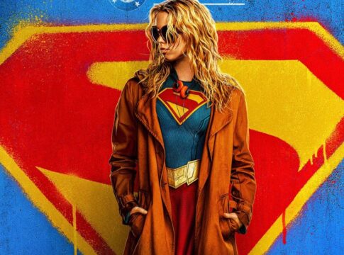Foto promocional do filme Supergirl onde a atriz Milly Alcock (mulher loira, de óculos escuros e casaco preto por cima do uniforme de super-heroi) aparece olhando para o lado