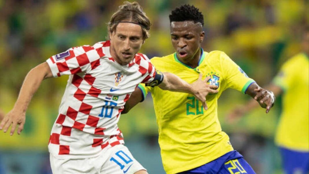 Jogador da seleção croata, com uniforme branco e vermelho quadriculado, disputa a bola com um jogador da seleção brasileira, que veste camisa amarela e shorts azuis; ambos correm lado a lado em campo, com outros atletas desfocados ao fundo