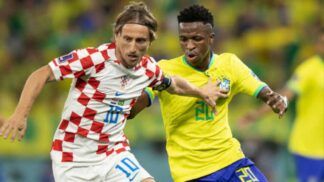 Jogador da seleção croata, com uniforme branco e vermelho quadriculado, disputa a bola com um jogador da seleção brasileira, que veste camisa amarela e shorts azuis; ambos correm lado a lado em campo, com outros atletas desfocados ao fundo