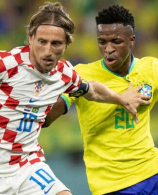 Jogador da seleção croata, com uniforme branco e vermelho quadriculado, disputa a bola com um jogador da seleção brasileira, que veste camisa amarela e shorts azuis; ambos correm lado a lado em campo, com outros atletas desfocados ao fundo
