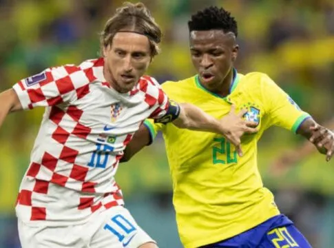 Jogador da seleção croata, com uniforme branco e vermelho quadriculado, disputa a bola com um jogador da seleção brasileira, que veste camisa amarela e shorts azuis; ambos correm lado a lado em campo, com outros atletas desfocados ao fundo