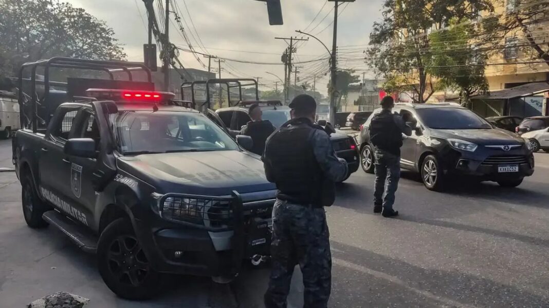 Policiais militares, de costas, carregam armas ao lado de camburões
