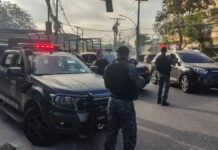 Policiais militares, de costas, carregam armas ao lado de camburões