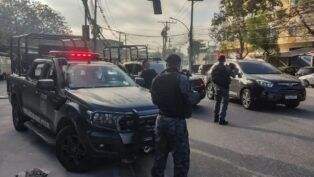 Policiais militares, de costas, carregam armas ao lado de camburões