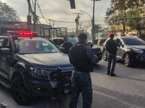 Policiais militares, de costas, carregam armas ao lado de camburões