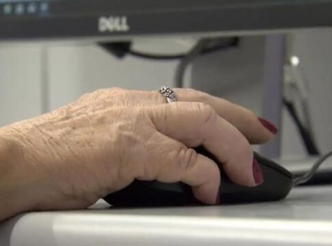 Uma mão de uma mulher idosa mexe no mouse diante de um computador