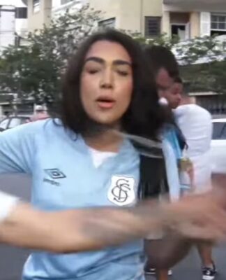 Mulher de camiseta azul-pastel do Santos surpreendida por um homem - na imagem, um borrão, pois estava em rápido movimento - de boné e vestindo camiseta branca do Santos. O homem toma o microfone da mão da repórter.