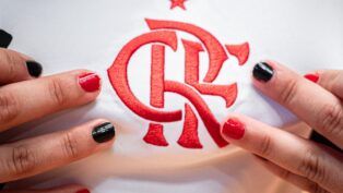 Close no escudo do Flamengo em uma camisa branca de jogo. Perto do escudo estão dedos com unhas pintadas de preto ou vermelho