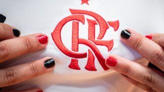 Close no escudo do Flamengo em uma camisa branca de jogo. Perto do escudo estão dedos com unhas pintadas de preto ou vermelho